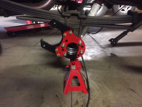 1982 Ford Bronco - A close-up view of a red and black suspension part, possibly a brake caliper bracket, mounted on a red jack stand beneath a vehicle. The pa…