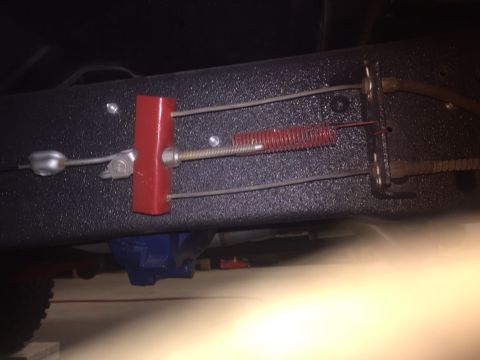 1982 Ford Bronco - A close-up view of the parking brake cable assembly mounted to the frame of a vehicle. The assembly includes a red bracket, a coiled red sp…