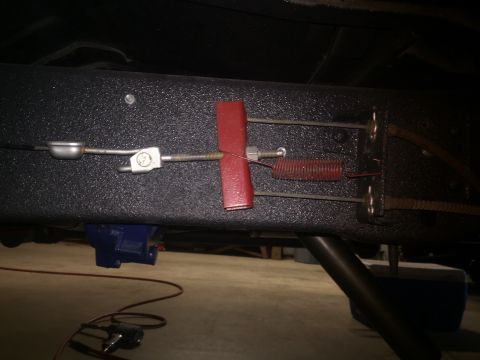 1982 Ford Bronco - A close-up view of the undercarriage of a vehicle, focusing on the parking brake cable mechanism attached to the frame. A blue actuator is…
