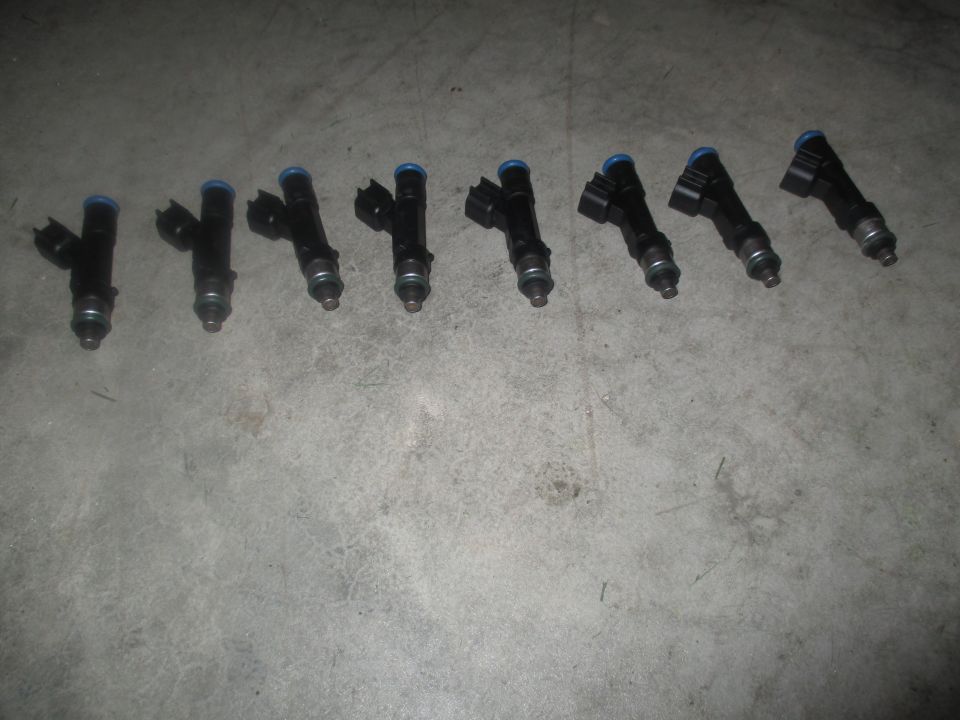 Eight black fuel injectors with blue tops on a concrete surface.