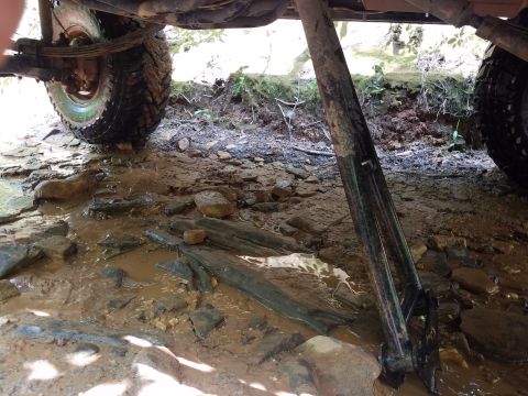 1988 Ford Bronco - The underside of a 1988 Ford Bronco is shown, with the rear tire resting on muddy ground near a stream. A Hi-Lift jack is positioned under…