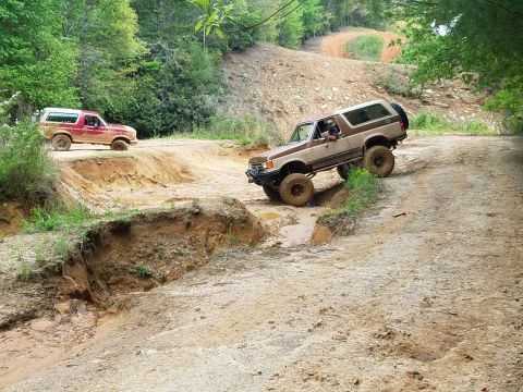 1988 Ford Bronco - Two off-road SUVs navigate a muddy, rutted trail with steep inclines and water obstacles. The lead vehicle, a tan SUV, is angled sharply as…