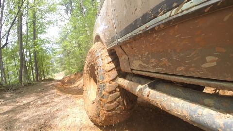 1988 Ford Bronco - A close-up, low-angle shot of the muddy tire and lower body of an off-road vehicle on a dirt path through a forest.