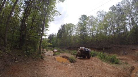 1988 Ford Bronco - A lifted, dark-colored Ford Bronco is navigating a muddy off-road trail, with another vehicle visible further up the path. The scene is set…