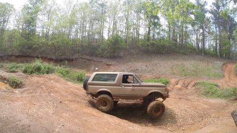 1988 Ford Bronco - A tan and brown Ford Bronco is shown off-roading on a dirt trail with mud and puddles. The vehicle is angled to the side, with its front wh…