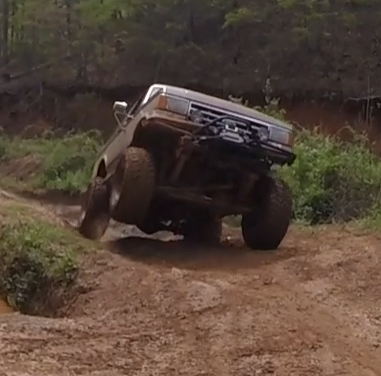 1988 Ford Bronco - A lifted, mud-covered pickup truck is angled sharply to the right, with its front passenger-side wheel high in the air, navigating a muddy…