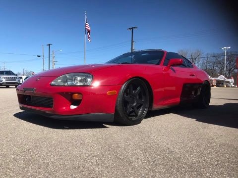 1994 Toyota Supra - A red Toyota Supra is parked on asphalt under a clear blue sky with an American flag in the background.