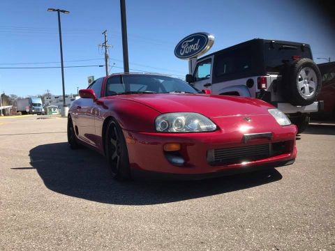 1994 Toyota Supra - A red 1994 Toyota Supra is parked outside a dealership, with a Jeep Wrangler visible in the background.