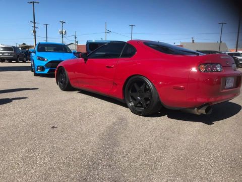 1994 Toyota Supra - A red sports coupe is parked in a lot next to a blue hatchback under a clear sky.