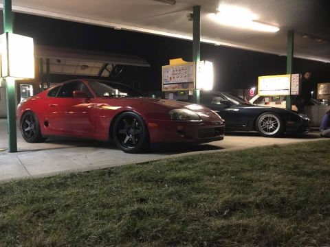 1994 Toyota Supra - Two sports cars, a red Supra and a black car, are parked under a drive-thru canopy at night.
