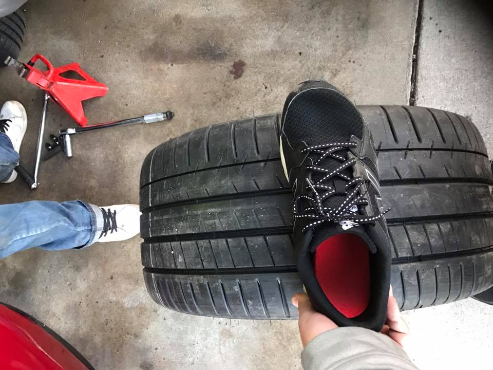 Two tires stacked, with a black sneaker placed on top. A red jack and lug wrench are visible in the background