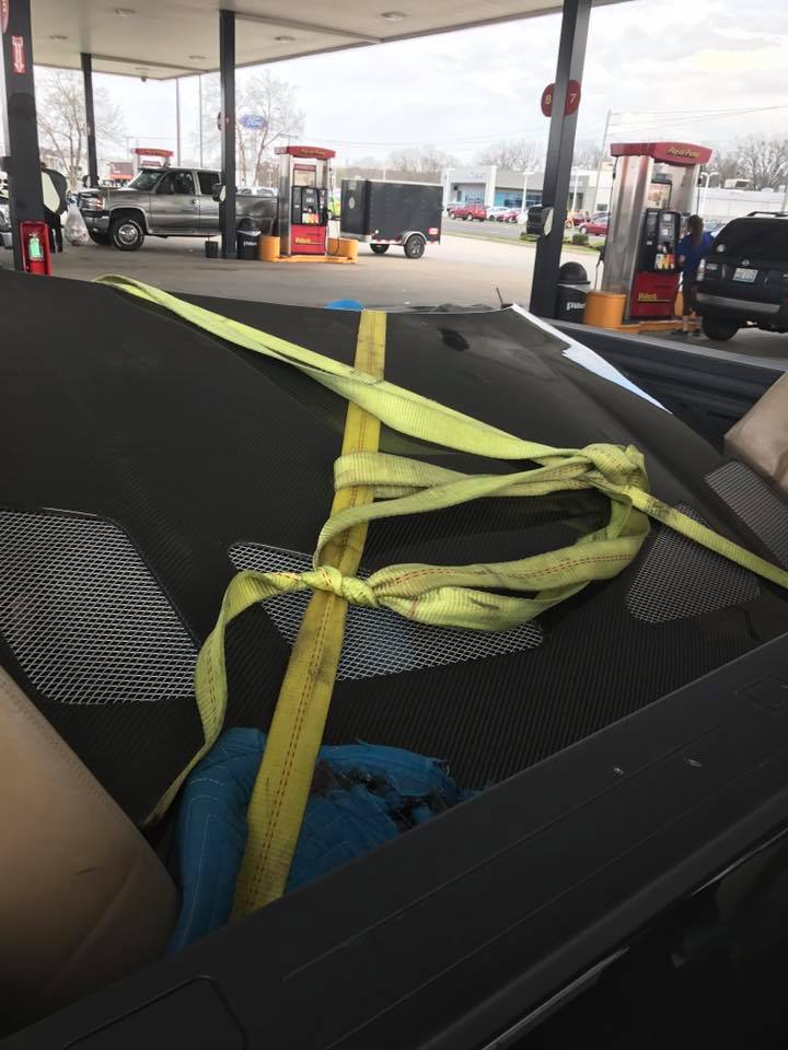 Car hood tied down with yellow straps at a gas station. Pickup truck and trailer visible
