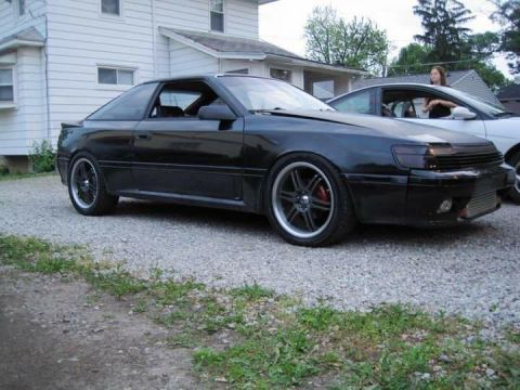1988 Toyota Celica - A black coupe with custom modifications is parked in a gravel driveway next to another vehicle.