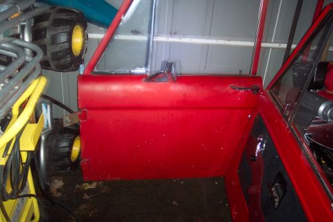 1974 Ford Bronco - Red vehicle door and interior visible, with large tires and yellow equipment in background.