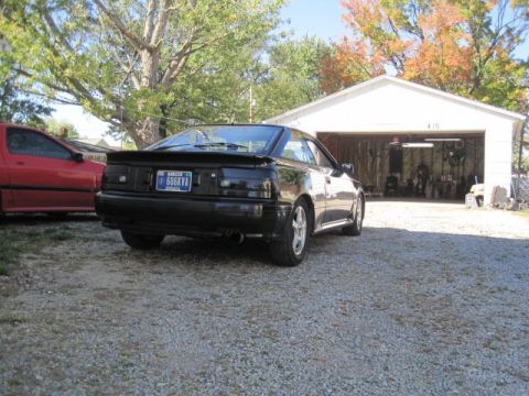 1988 Toyota Celica - A black coupe with a spoiler is parked in a gravel driveway next to a red car, with a garage in the background.