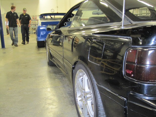 Black car side view with aftermarket wheels and tinted taillights. Another blue car is visible behind it