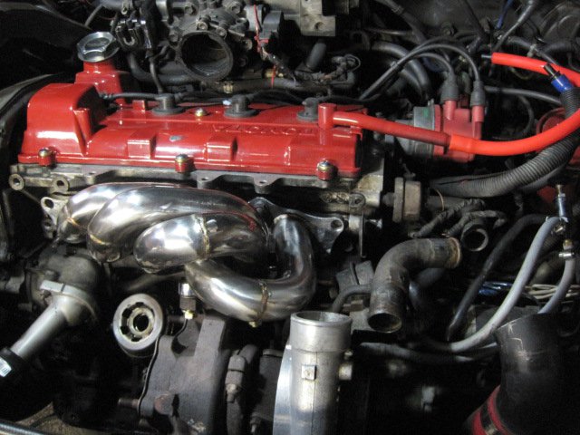 Engine bay showing a red valve cover and a shiny custom exhaust manifold. Various hoses and wires are visible
