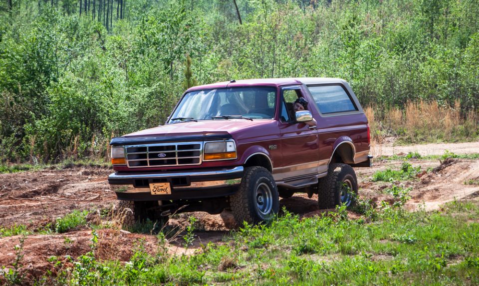 Maroon Ford Bronco with tan trim driving on a dirt path, front-left view