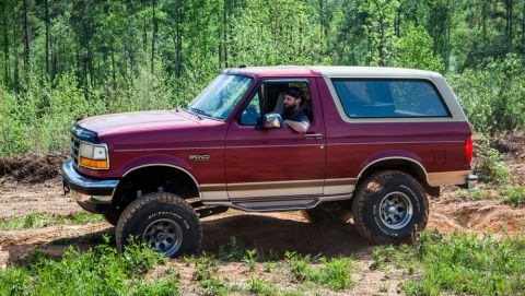1995 Ford Bronco - 1995 Ford Bronco off-roading on a dirt trail surrounded by trees.