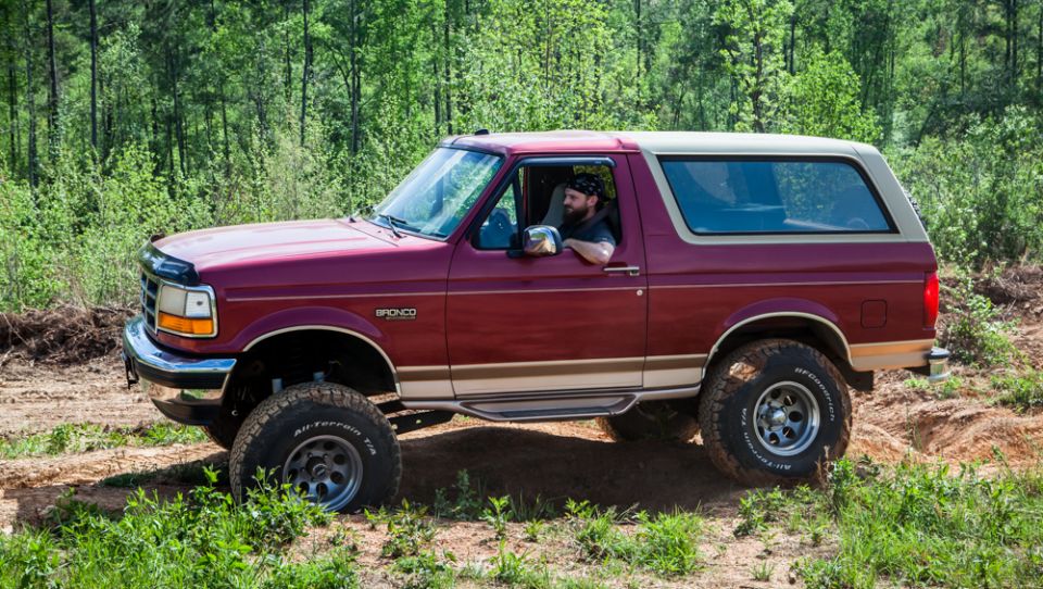 Maroon 1995 Ford Bronco with tan trim driving on a dirt path in a wooded area