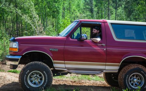 1995 Ford Bronco - A man drives a red Ford Bronco off-road through a wooded area.