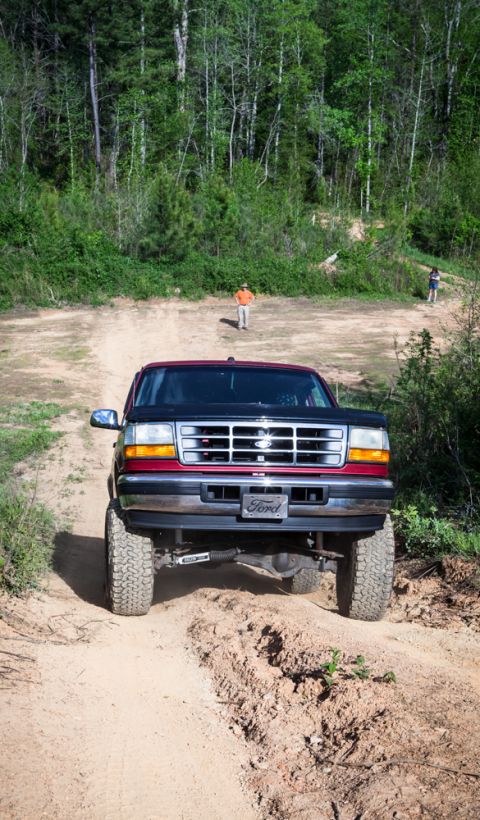 1995 Ford Bronco - A Ford truck with a prominent grille and lifted suspension navigates a dirt path through a wooded area.