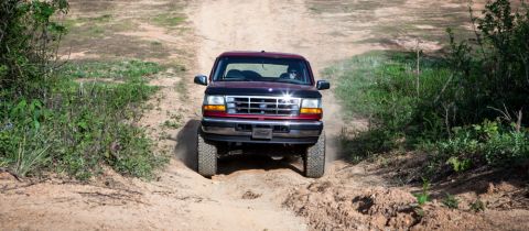 1995 Ford Bronco - A maroon Ford Bronco navigates an off-road trail, kicking up dust as it ascends a sandy incline.