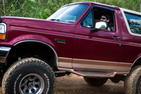 1995 Ford Bronco - A maroon 1995 Ford Bronco is seen from the front-three-quarter angle, with the driver visible behind the wheel.