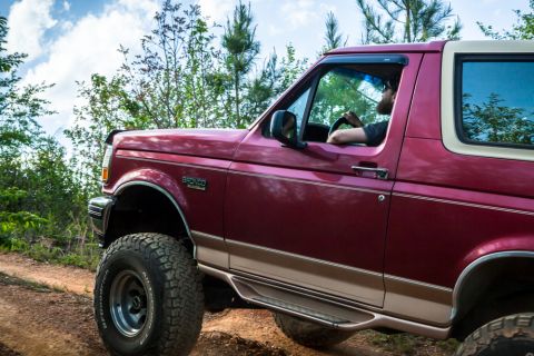 1995 Ford Bronco - A 1995 Ford Bronco is driven off-road on a sunny day with a blue sky and clouds.