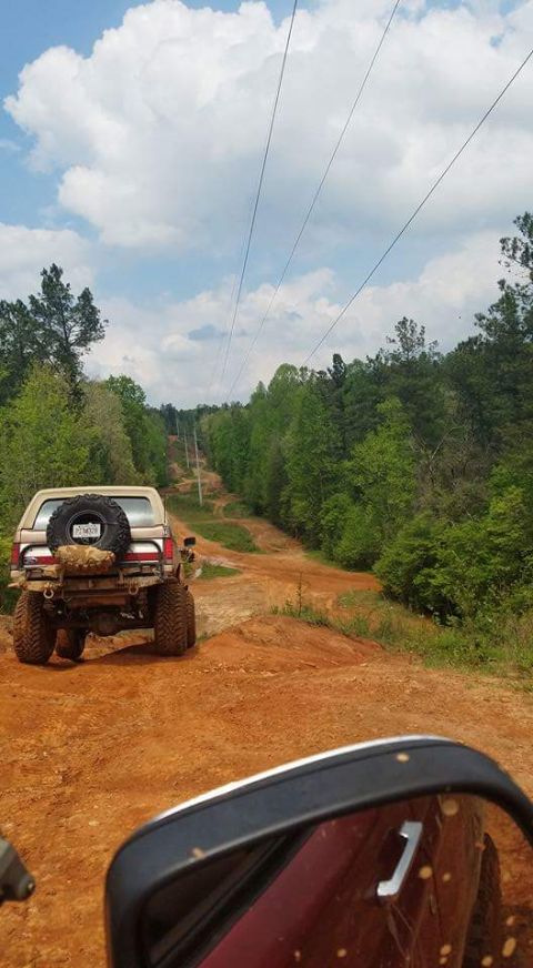 1988 Ford Bronco - A lifted Ford Bronco navigates a dirt path through a wooded area under a cloudy sky.