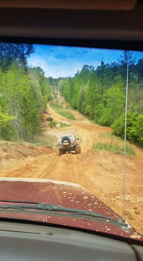 1988 Ford Bronco - An SUV navigates a muddy, off-road trail surrounded by trees, with another vehicle ahead on the path.
