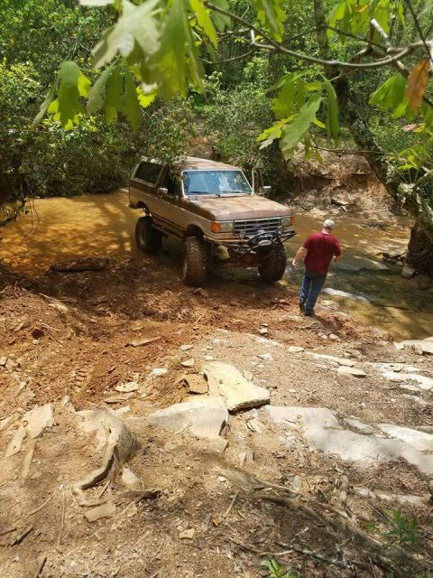 1988 Ford Bronco - A man walks near a lifted Ford Bronco as it drives through a wooded area with a muddy stream.