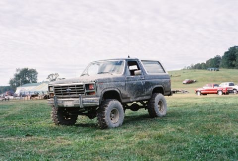 1986 Ford Bronco - A muddy Ford Bronco sits in a field with other vehicles visible in the background under a cloudy sky.