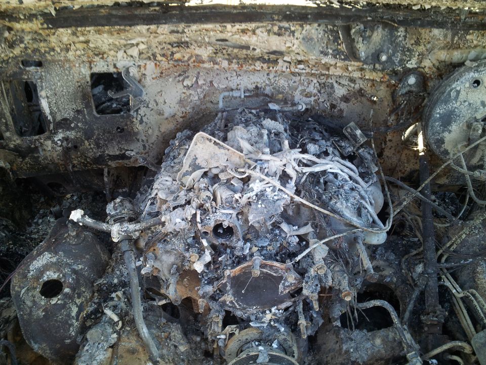 Fire-destroyed engine bay with burned engine, melted wiring, and rusted charred metal
