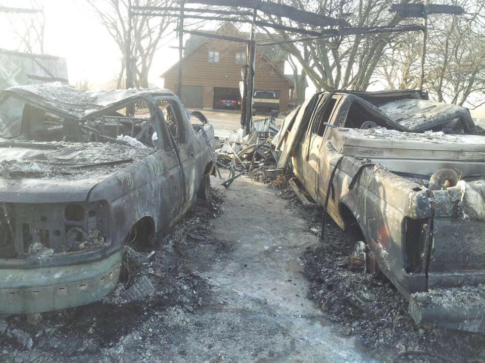 Two burned-out trucks reduced to charred shells amid ash and debris, with a log house in the background