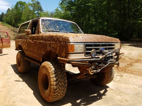1988 Ford Bronco - A heavily mud-covered tan Ford Bronco is parked on a dirt path surrounded by trees. The off-road vehicle features large tires, a winch on t…