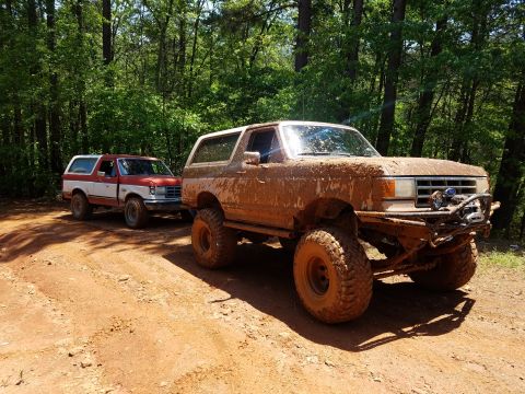 1988 Ford Bronco - Two heavily muddied Ford Broncos are parked on a dirt trail surrounded by trees. The Bronco in the foreground is a dark color and has a win…
