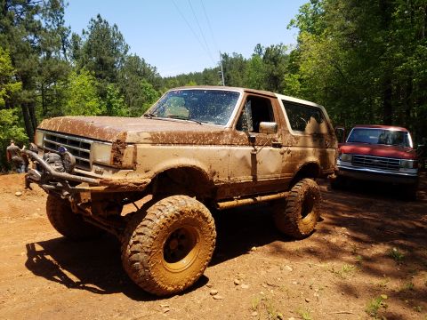 1988 Ford Bronco - A heavily mud-covered, lifted 1980s Ford Bronco is parked on a dirt path in a wooded area, with a red Ford pickup truck visible behind it.