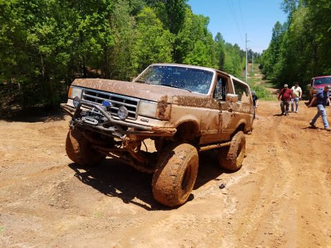 1988 Ford Bronco - A mud-covered 1980s Ford Bronco is parked on a dirt road surrounded by trees. Several people are visible in the background near another veh…
