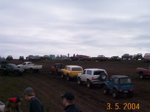 1983 Ford Bronco - A line of off-road vehicles, including a white 1983 Ford Bronco, navigate a muddy terrain during an event.