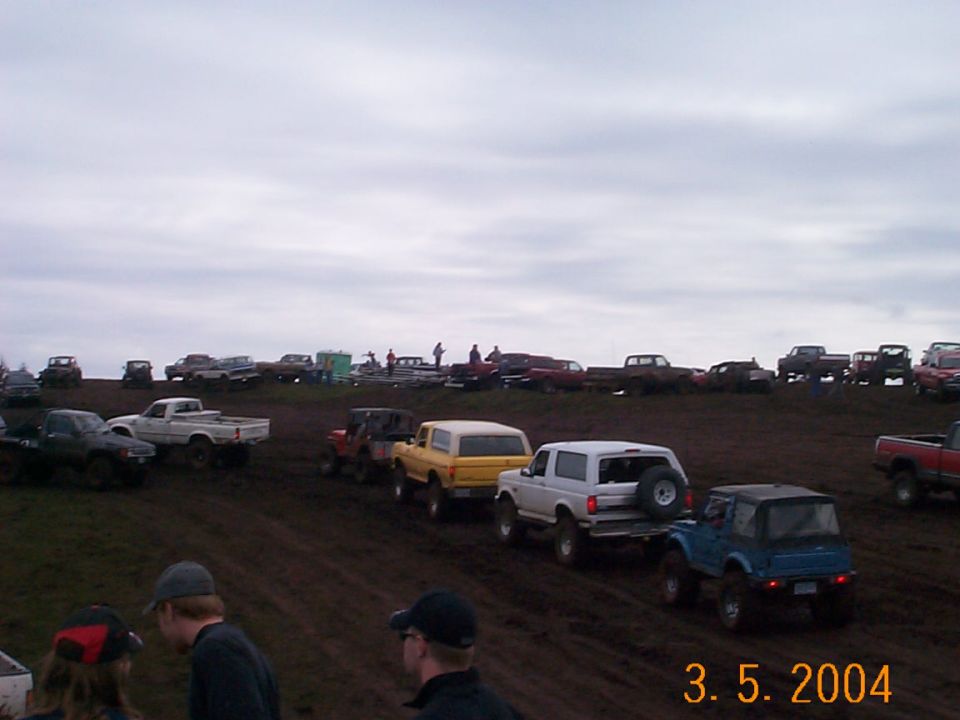 Row of 4x4 trucks and SUVs lined up in the mud at an off-road event, spectators watching