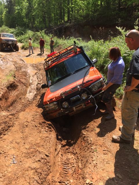 1988 Ford Bronco - An orange off-road vehicle is stuck in a deep mud rut on a dirt trail, with several people standing around it. Another tan off-road vehicle…