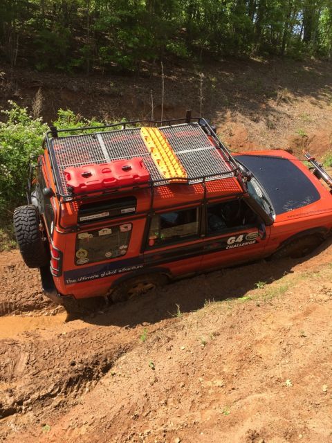 1988 Ford Bronco - An orange off-road vehicle is stuck in thick mud, with its tires sunk deep into the mire. The vehicle is equipped with a roof rack carrying…