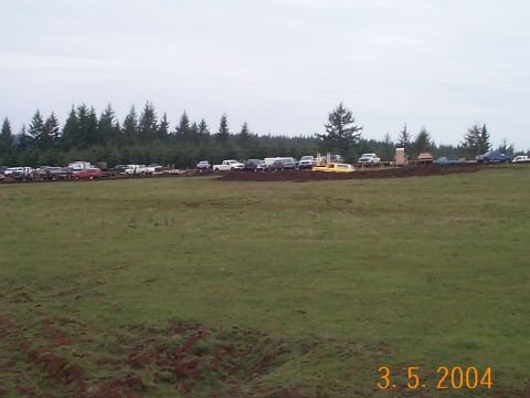 1983 Ford Bronco - A line of vehicles are parked in a field with trees behind them under a cloudy sky.