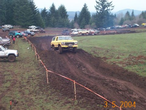 1983 Ford Bronco - A yellow SUV navigates a challenging muddy course with spectators and other vehicles in the background.
