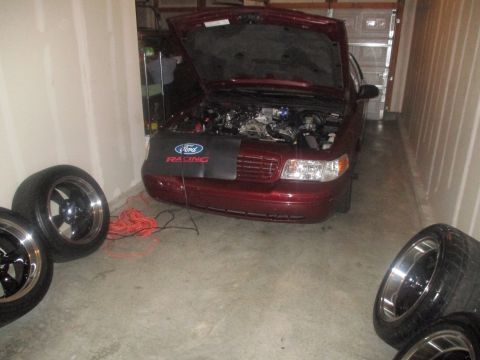 2004 Ford Crown Victoria - A maroon Ford Crown Victoria with its hood open is parked in a garage, showing the engine bay. Several aftermarket wheels and a coil of ora…
