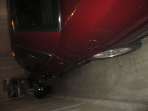 2004 Ford Crown Victoria - The underside of a dark red sedan is visible, with a portion of the rear wheel and tire showing. A metal ramp or lift is positioned beneath…