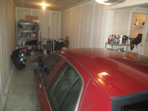 2004 Ford Crown Victoria - A red sedan is parked in a garage with its side facing the camera. Various items are stored on shelves and on the floor in the background.