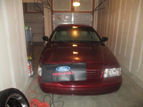 2004 Ford Crown Victoria - A maroon Ford Crown Victoria is parked in a garage, facing forward. A black car bra with a Ford Racing logo is covering the front grille.