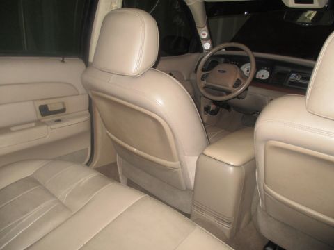 2004 Ford Crown Victoria - The interior of a tan Ford Crown Victoria is shown, focusing on the front seats, center console, and dashboard. The driver's side door pane…
